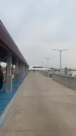 The video presents a view of a pier on an overcast day. People walk along the concrete walkway toward a large, white boat docked at the end. A covered walkway with a brown roof and gray pillars runs along the left side of the concrete walkway. On the right, a gray concrete barrier separates the walkway from the water. The camera pans slightly to the right, showing a small boat docked next to the pier. Further out, a long rock barrier stretches across the water, and distant land with trees is visible on the horizon. The overall atmosphere appears calm and quiet.