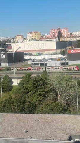 The video shows a cityscape under a clear blue sky. In the foreground, a highway runs horizontally across the frame, partially obscured by trees and greenery. A train passes by on the highway. Behind the highway, there are buildings, including a large white building with the name "DYRUP" written on it, a black building labeled "hub 361", and a red building with the text "VIVA". The buildings are interspersed with greenery and parked cars. A crane is visible in the background. The video is shot from an elevated position, looking down at the scene. The overall impression is a bright, sunny day in an urban environment.