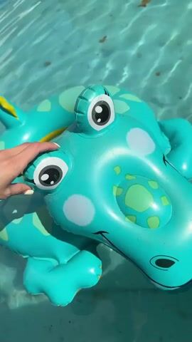 The video shows a child's inflatable crocodile floaty floating in a round above-ground swimming pool, along with a smaller inflatable boat. The crocodile floaty is aqua-colored with light green and yellow accents, white polka dots, and cartoonish eyes. A hand briefly touches the floaty before it drifts in the water. The pool has a light blue liner, and some debris is visible on the bottom. The surrounding environment shows grass and some items scattered in a yard.