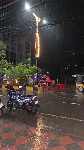 The video captures a nighttime street scene during rainfall. A motorcycle is parked on the left side of the frame, on a checkered patterned sidewalk that is wet from the rain. Across the street, there are several vehicles including auto-rickshaws and bicycles, some of which appear to be in motion, creating motion blur. People are also visible, some under umbrellas. A street vendor stall is present on the right side of the road. A decorative light fixture hangs from a pole, illuminated by a warm light. The street and sidewalk are wet, reflecting the lights from the street lamps and vehicles. The overall atmosphere is that of a busy urban street during a rainy night.