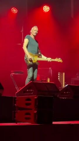 A man, identified as Sting, stands on a stage playing a bass guitar. He is wearing a dark t-shirt and jeans. The stage is lit with red and purple lights, and there are several speakers stacked on the stage. He appears to be performing for an audience, though the audience is not visible in the frame. The lighting changes from red to purple throughout the short clip.