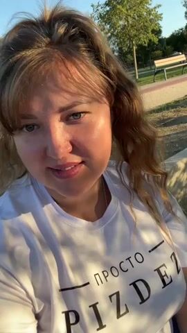 A woman with light brown hair pulled into a high ponytail, bangs, and green eyes is captured in a close-up selfie-style video. She is wearing a white t-shirt with black text that reads "ПРОСТО PIZDE". The video is taken outdoors in what appears to be a park, with trees and a bench visible in the background. Sunlight casts shadows on the woman's face, highlighting her features. She smiles and tilts her head slightly, interacting with the camera.