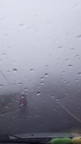 The view from inside a vehicle driving on a wet road with low visibility due to fog and rain. Raindrops are visible on the windshield. A motorcycle is in front of the vehicle, traveling in the same direction. A streetlight is visible on the side of the road. Another vehicle appears briefly in the opposite lane. The overall atmosphere is foggy and wet.