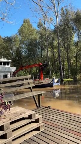The video opens with a static shot of a waterway alongside a wooden dock, with a boat equipped with an orange crane visible on the water. The boat, named "Tango," appears to be stationary. Trees and a clear blue sky form the backdrop. A wooden picnic table is situated next to the dock. The water is a murky brown. The camera then shifts to a close-up view of a mate cup filled with yerba mate and a small space for adding hot water. Parts of the person holding the mate are visible, including white pants, a green thermos, a small potted plant, and one black shoe.