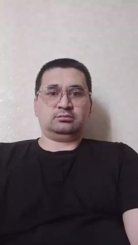 A man with dark hair and glasses is looking directly at the camera. He is wearing a black t-shirt. The background is a textured wall.
