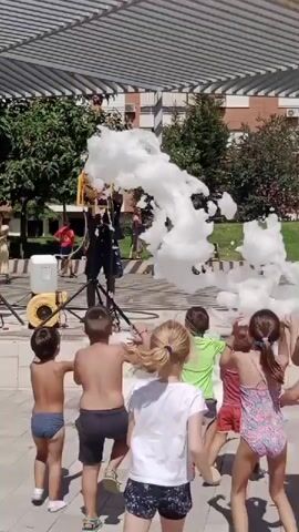 This video captures a sunny outdoor event, likely a children's party or festival, centered around a foam machine. Several children, mostly in swimwear, are actively engaged with the copious amounts of white foam being generated by a large machine on the left side of the frame. The foam machine itself appears to be a portable unit with a yellow tank and a fan. A person, possibly an entertainer, stands behind the machine, seemingly controlling the foam output. The children are positioned in front of the machine, with some reaching out to touch or play in the foam, while others observe with excitement. In the background, there's a shaded area with a white canopy, trees, and buildings, suggesting an urban or suburban park setting. The ground is paved, and the overall atmosphere is one of joyful activity and summer fun.