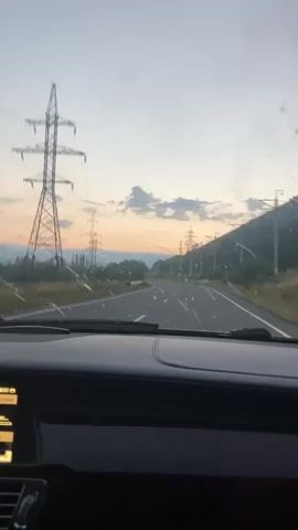 This is a point-of-view video from inside a car, looking out the windshield. The car is driving on a paved road with white lines marking the lanes. The road curves slightly to the left in the distance. The sky is a pale blue with some wispy clouds, and it appears to be either sunrise or sunset, with a faint orange glow on the horizon. There are several large, tall electrical towers with wires visible along the side of the road. The landscape is hilly, with trees and vegetation on the slopes. The dashboard of the car is visible in the foreground, and the windshield has some smudges or water droplets on it. There are no people visible in the video, and no other vehicles are close by. The overall impression is one of traveling through a rural area during twilight.