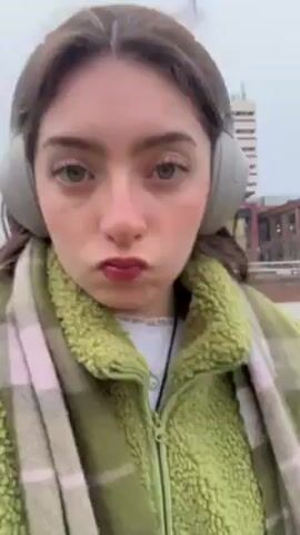A young woman with dark hair and fair skin is walking outside and recording a selfie video. She has headphones on and is wearing a green fleece jacket, a plaid scarf, and a light blue purse. She is making a pouting face at the camera. The background includes buildings, a sidewalk, and some greenery. The weather appears to be overcast.