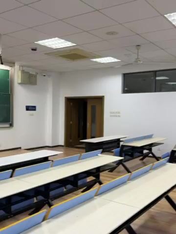 The video depicts a static shot of an empty classroom. It shows rows of desks with blue dividers and black legs. The floor is wooden. The walls are white, and there's a rectangular window near the ceiling on one wall. The ceiling is white with square light fixtures. A ceiling fan is also visible. A door is present on one of the walls. There are no people in the scene.