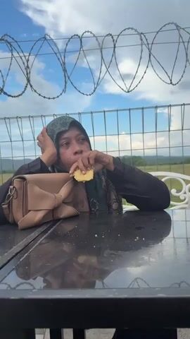 A video begins with a woman wearing a hijab and black clothing sitting at a table outdoors, holding a piece of food up to her mouth. A light brown handbag is on the table next to her. Behind her is a barbed wire fence and a cloudy sky. The camera pans slightly to the right, showing another woman with a light purple hijab and glasses sitting at a table with a white purse and a phone. The scene then shifts to show a wider view of an outdoor seating area with several tables and chairs. People are seated at some of the tables. In the background, there are more fences, fields, and buildings. The camera continues to pan right, revealing more tables and chairs, some potted plants with red flowers, and a streetlamp. The video ends with a close-up of a hand holding a piece of food.