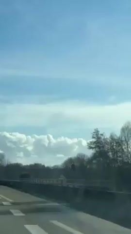 The video shows a view from inside a moving vehicle, likely a car, traveling on a highway. The sky is blue with scattered white clouds. There is a barrier beside the highway, and beyond that, a field and trees. A sign indicating a speed limit of 110 can be seen beside the road. The day appears to be sunny.