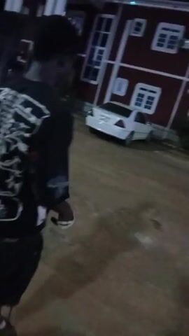 A person is standing outdoors on a paved area at night. The person is wearing a black t-shirt with white graphics on the back and front, black shorts, and flip-flops. They are holding something in their hands, possibly looking at it. In the background, there is a white car and a red and white building. A street light is visible, illuminating the scene.