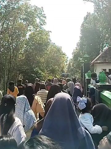 A large group of people are gathered outdoors on a sunny day, many wearing headscarves. The crowd is dense, and the view is mostly of the backs of people's heads and shoulders. In the background, trees line a path or road. Some people appear to be looking towards a structure or area further down the path. There are also what appear to be decorative green objects and a building visible on the right side of the frame. Some children are present in the crowd. The overall impression is of a public gathering or event.