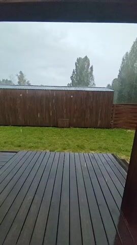 This video shows a view from a wooden deck, looking out at a backyard during a rain shower. The deck boards are wet and dark. In the background, there is a section of green grass, a tall, dark wooden fence, and trees. The sky is overcast and grey, with visible raindrops falling. There is a small wooden structure on the grass in front of the fence. The overall scene is one of a dreary, rainy day.