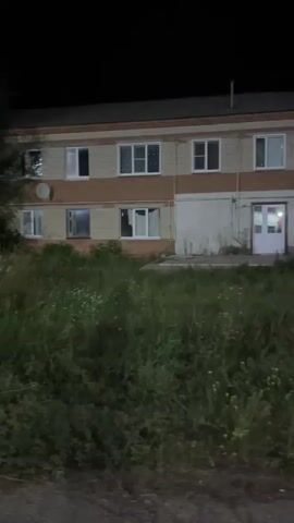The video shows a dimly lit, two-story brick building at night. The building has several windows, some with lights on inside. There is an entrance with a white door and an air conditioner unit above it. In front of the building, there is an area with tall, overgrown grass and some scattered plants. A large bush or small tree is visible on the right side of the frame, partially obscuring the building. The ground in the foreground appears to be a dirt or gravel path. The sky is dark, indicating nighttime. The camera pans slightly to the right, revealing more of the building and the surrounding overgrown vegetation.