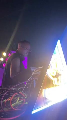 A DJ is seen from the chest up, wearing headphones and glasses, operating a DJ mixer. The DJ booth is illuminated by purple and yellow stage lights. Wires from the equipment are visible. A crowd is out of focus in the background, with some individuals wearing striped shirts. The environment appears to be a nightclub or a similar venue with low lighting and vibrant stage effects.