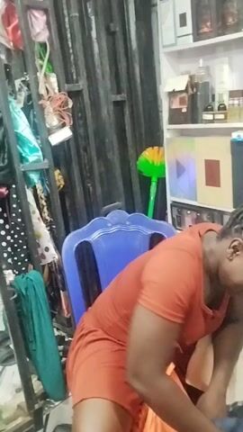 A woman in an orange dress is seen kneeling down and rummaging through a black trash bag. She is surrounded by shelves filled with various items, including perfumes, toiletries, and other products. The environment appears to be a shop or a storage area. The woman is focused on the contents of the trash bag, and her actions suggest she is searching for something or sorting through items.
