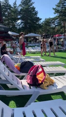A wide shot shows a sunny day at an outdoor recreational area with a pool and lounge chairs. In the foreground, several white lounge chairs are visible, one with a pink backpack and a yellow towel on it. Beyond the chairs, a bright green artificial turf area is populated with people. In the middle ground, a group of people, including children and adults in swimwear, are gathered near the edge of a blue swimming pool. Some are sitting on the grass, while others are in the water. In the background, a line of white umbrellas shades more lounge chairs. Tall pine trees form a dense forest surrounding the area. The sky is clear and blue.
