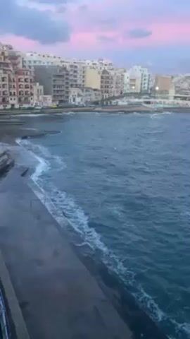 The video shows a coastal scene with waves lapping against a concrete embankment. In the background, there are several buildings of varying heights and colors. The sky is a blend of pink and gray, suggesting either a sunrise or sunset. The water is a deep blue-green, with white foam visible where the waves break. The video is relatively still, providing a stable view of the scene.