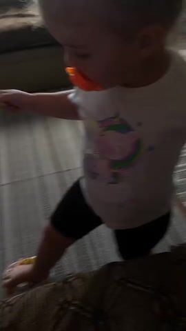 A toddler wearing a white t-shirt with a unicorn design and black shorts with an orange pacifier in their mouth is playing on the floor. The toddler has their left foot on a small, multi-colored ball, and their right hand is resting on a patterned cushion. Another person's leg wearing white socks is visible in the lower left of the frame. The floor is covered with a striped rug. In the background, a sectional sofa is partially visible.