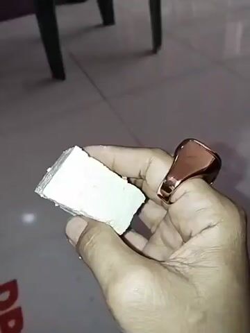 The video shows a hand holding a piece of white cheese. The cheese is a rectangular block, and the hand is holding it in a way that it can be examined from different angles. The hand is wearing a ring on the index finger. The background is blurred, but it appears to be an indoor setting with a tiled floor and furniture. The lighting is somewhat dim. The video appears to be a short clip, possibly a demonstration or a review of the cheese.