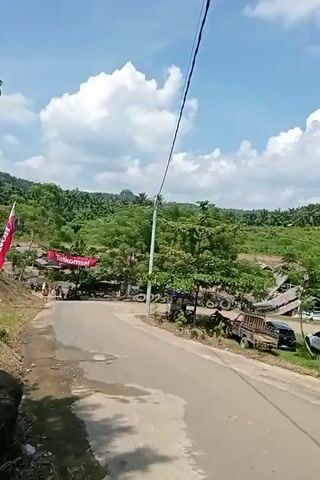 The video pans across a rural scene on a sunny day with fluffy white clouds. In the foreground, a paved road curves to the right. To the left of the road, there is a red banner that reads "Telkomsel." Beyond the banner and road, there are trees and a few wooden structures with thatched roofs, resembling stalls or small buildings. Several vehicles are parked in the area, including trucks and cars. Further in the background, the landscape is covered in lush green trees and vegetation, suggesting a hilly or mountainous terrain. The overall impression is of a community gathering or a roadside market in a natural setting.