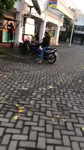 A woman wearing striped pants sits on a motorcycle parked on a brick street. She is facing away from the camera, looking toward a food stand in the background. The food stand is manned by a person wearing a maroon shirt and a black head covering, who is preparing food. There is another motorcycle parked behind the woman's. The street is lined with buildings.