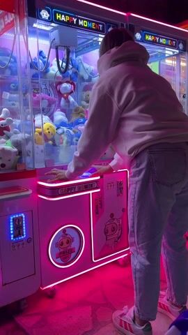 A person wearing a white hooded sweatshirt, light blue jeans, and white and red Adidas sneakers is playing a claw machine arcade game. The claw machine, labeled "HAPPY MOMENT," is illuminated with bright neon pink and blue lights, emphasizing its playful design and the array of plush toys inside. The person is focused, using the controls to position the claw over the stuffed animals. The arcade is bathed in a vibrant pink hue. The arcade game is designed with a cute robot character and circular "PRIZE OUT" label.