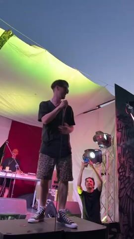 A male performer wearing sunglasses, a black t-shirt, and leopard-print shorts stands on a stage holding a microphone. He is facing left and smiling. Behind him, another male is seated at a DJ setup. To the right of the performer, a third male is holding up a phone, seemingly recording. The stage is covered by a large white tarp illuminated with green and pink lights. The background appears to be an outdoor setting with a clear blue sky.