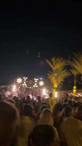 A large crowd of people are gathered outdoors at night, seemingly at a party or event. The sky is dark, with some lights illuminating the area. In the background, there is a structure with lights hanging from it, and palm trees are visible on the right side of the frame, lit up from below. The people in the foreground are facing towards the background, with many holding up their phones, suggesting they are watching or recording something. The overall atmosphere appears to be festive and energetic.
