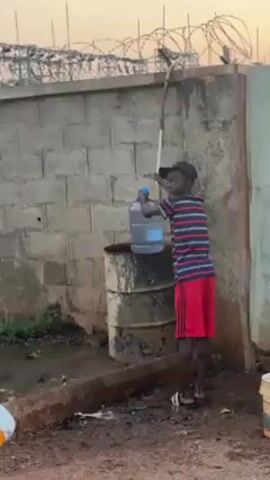 A boy is filling a large water jug at a water source in front of a cinder block wall. The boy wears a striped shirt, red shorts, a black cap and sandals. He leans against a long pole while filling the jug, which sits on top of a rusty barrel. The wall is topped with barbed wire. There is some greenery near the base of the wall, and the ground is wet. The sky is visible in the background.