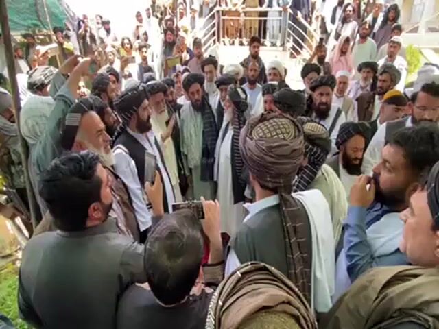 Certainly! Here's a detailed description of the video:

**Scene Overview:**

The video shows a large group of men clustered together in an outdoor setting. The men are primarily wearing traditional Afghan clothing, including turbans, vests, and long shirts. The scene seems to be taking place near or in front of a building with a modern architectural design.

**People:**

*   Most of the men are wearing turbans or headscarves, common attire in Afghanistan.
*   Some men are dressed in more modern attire, such as collared shirts or blazers, indicating a possible range of social status or roles.
*   A man is recording on a phone

**Actions & Interactions:**

*   The crowd is densely packed, with many individuals jostling and moving slightly.
*   There are some interactions between individuals, with some turning to speak to one another.

**Environment:**

*   The setting appears to be outdoors, possibly in a courtyard or open area near a building.
*   The architecture in the background looks relatively modern.
*   The lighting suggests it is daytime.