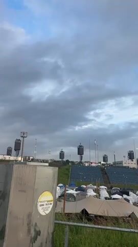 A low-angle, vertical, outdoor, right-to-left pan shows a parking lot with a tent-filled camping ground and a stadium beyond a metal railing. The camera pans from left to right, revealing several black and white tents, a large white tent, and another section of the stadium. The sky is cloudy with hints of blue.