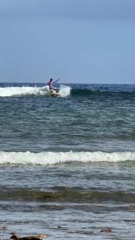 A surfer in a light purple long-sleeved shirt rides a wave in the ocean under a bright blue sky. The surfer is standing on a white surfboard, arms outstretched for balance. The ocean is a deep blue-green with small white-capped waves rolling towards the rocky shoreline in the foreground. The camera is stationary, providing a steady shot of the surfer's progress.