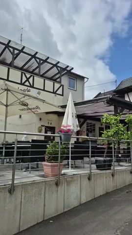 The video presents a street scene in what appears to be a European village. The camera executes a slow pan to the right, then a slight pan back to the left. On the left side of the street, a two-story building features a white facade with dark timber framing on its upper floor, accompanied by a modern extension with a glass roof. An outdoor dining area with tables, chairs, potted plants, and a white umbrella is situated in front of this building, enclosed by a metal railing. On the right side of the street, another outdoor dining area with tables, chairs, potted plants, and a large maroon umbrella is visible, also bordered by a metal railing. A street sign, partially reading "Serva," is visible in the upper right. Further down the street, a building with a tall, pointed steeple, indicative of a church, is seen in the distance, with parked cars along the street. The sky is partly cloudy. No individuals are present in the video.