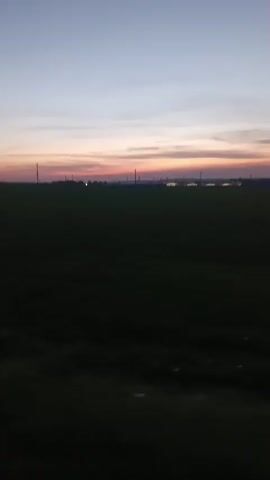 The video shows a dark field in the foreground with a distant horizon illuminated by a sunrise or sunset. The sky displays hues of blue, pink, and orange. In the distance, there are faint outlines of what appear to be industrial structures, possibly towers or pylons. The overall scene is static and peaceful.
