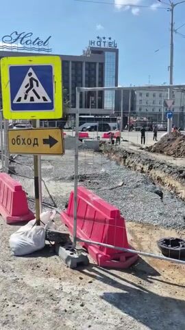 The video shows an outdoor construction site on a sunny day. In the foreground, there are two red plastic traffic barriers, a white plastic bag, and a white metal pole. Behind the barriers, a trench is visible, filled with gravel and a large concrete ring. To the left of the trench, a yellow sign with a pedestrian crossing symbol and an arrow indicating an "обход" (detour) is visible. In the background, a modern hotel building with "HOTEL" and three stars displayed on its facade stands tall. Several construction workers in orange vests are visible working on the site, and a bus and some cars are on the road behind the construction area. The overall environment appears to be a city street undergoing roadwork or utility installation.