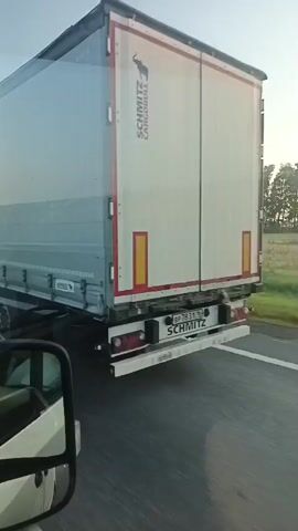 The video shows a side view of a large white truck with a grey tarp covering its cargo area. The truck is moving on a road, as evidenced by the motion blur and the presence of a road marking. A side mirror of another vehicle is visible in the foreground on the left. The truck has multiple axles and wheels. The environment appears to be outdoors, with a hint of trees and fields in the background. The sky is visible at the top of the frame. The video is shot from a moving vehicle, likely another truck or a car, traveling alongside the first truck. The camera is positioned to capture the side of the truck. The scene appears to be during daylight.