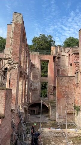 The video shows the interior of a partially ruined brick building. The walls are made of red brick and have sections of crumbling plaster, revealing the brickwork underneath. Large rectangular openings, once windows or doorways, are visible in the walls, looking out onto greenery. Scaffolding is erected in several areas, suggesting ongoing restoration or construction work. Two individuals, wearing hard hats and work attire, are visible in the foreground, seemingly engaged in activities related to the scaffolding or the building's structure. The ground appears to be a mixture of dirt and debris, with some wooden planks laid out. The sky above is blue with scattered white clouds. The overall impression is one of historical architecture undergoing preservation.