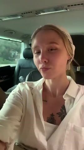 The video shows a young woman with blonde hair, wearing a white shirt and a bandana on her head. She has a tattoo on her chest and a nose ring. The woman is sitting in the passenger seat of a vehicle, likely a van or SUV. The interior of the vehicle is visible, including the headrest of the driver's seat and the ceiling. Through the windows, trees and foliage can be seen, suggesting an outdoor setting, possibly a rural or wooded area. The lighting suggests it might be daytime, with natural light filtering into the vehicle. The woman appears to be smiling and looking at the camera, possibly taking a selfie or recording a short video. The overall mood seems casual and relaxed, as if she is on a journey or outing. There is no visible text in the media.