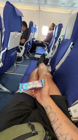 The video shows an interior view of an airplane cabin during a flight. The camera, held by a passenger, first focuses on their legs and a snack bar they are holding. The passenger is wearing black shorts and black socks. They have a tattoo on their arm. The video then pans to show other passengers seated in blue airplane seats, some of whom are looking out the window or sleeping. The cabin lighting is visible overhead. The view shifts to a wider shot of the airplane cabin, showing rows of blue seats and passengers. Finally, the camera captures a view of the airplane's wing from the window, against a bright, hazy sky. The overall impression is a typical airplane journey experience.
