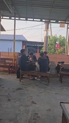 Four young males are gathered around a wooden table, seated on a wooden bench, under a corrugated metal roof. They appear to be engaged in a casual conversation or activity. The environment suggests an outdoor seating area, possibly a warung or cafe, with concrete flooring and some seating areas visible in the background. The lighting indicates it is daytime, possibly late afternoon, with the sun setting. The males are all wearing dark t-shirts. Two of them are looking down at the table, while the other two are looking towards each other. There is a backpack on the ground next to one of the males. The overall scene depicts a relaxed, social gathering.