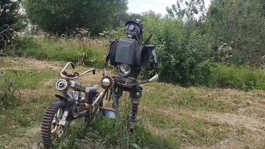 The video pans from left to right across a grassy outdoor area with trees and bushes in the background. In the foreground, on the left, is a sculpture of a robot made from scrap metal, standing next to a scrap metal motorcycle. The robot is holding a white object in its right hand. The camera continues to pan right, revealing a large, colorful sculpture of the Earth, also made from scrap materials, being held up by what appears to be a stylized insect. To the right of the Earth sculpture is a large padlock sculpture, and further right, a sunflower sculpture. The overall environment is a rustic, outdoor display of recycled art.