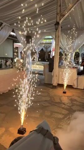 This video captures a wedding reception in an outdoor covered area. Two cold pyrotechnic fountains are active on the stone floor, emitting sparks and smoke. A bridal couple, the bride in a white wedding dress with a veil and the groom in a green suit, are dancing in front of the fountains. In the background, guests are seated at tables draped in white tablecloths and decorated with pink accents. The area is illuminated by string lights draped from the ceiling. A person is seen holding a phone, seemingly filming the couple.