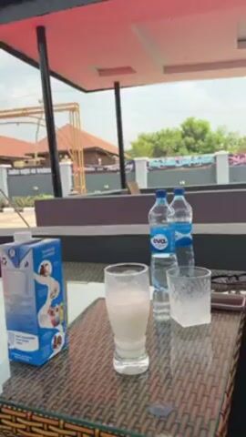 The camera is static, providing a view of a table on an outdoor patio. A carton of yogurt drink is on the left edge of the table. Next to it is a glass that is about two-thirds full of a white liquid. Two bottles of Eva water are placed near a small glass containing ice cubes. A pair of sunglasses rests next to the glass of ice. The table is made of a patterned wicker material with a glass top. The patio is covered by a red and white roof structure. In the background, other buildings, some foliage, and a cloudy sky are visible.