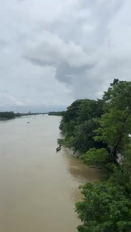 A wide, muddy river flows under a cloudy sky. The left bank is lined with dense green trees. On the right bank, a narrow strip of land is visible, with a small boat pulled up to the shore. Further in the distance, a bridge spans the river. The camera pans down to show a section of a white railing, indicating the vantage point is from a bridge or elevated structure. The water appears choppy in some areas, suggesting a current. There are no people visible in the immediate foreground or on the banks, though a figure can be seen in the small boat on the shore.
