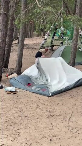A person with red hair and a tan shirt and black shorts is partially visible, hunched over and appearing to be interacting with a collapsed tent. The tent, which is white and gray, is lying on its side on a sandy surface. Behind the tent, there are several other tents set up in a wooded area with pine trees. Further in the background, other tents and picnic tables are visible. The scene transitions to a wider view of a beach with the ocean in the distance, under a cloudy sky. Several pieces of litter, including a plastic bottle, are scattered on the sand.
