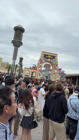 The video captures a very crowded outdoor scene, likely at the entrance of a theme park. A large archway, decorated with colorful balloons and a prominent "25th" emblem, is visible in the background, suggesting an anniversary celebration. Beyond the archway, multiple entrance gates can be seen, leading to a sprawling complex with a roller coaster structure rising high into the sky.

Hundreds of people, mostly young adults, are gathered in a dense crowd, standing and slowly moving forward. They are casually dressed, and some are seen talking or looking around. The atmosphere appears busy but orderly, with people seemingly queuing to enter the park or an attraction. The weather is overcast, suggesting a cloudy day. Based on the visual cues like the "25th" and "UNIVERSAL STUDIOS" partially visible on the archway, and the general appearance, the location is Universal Studios Japan in Osaka.