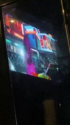 A person is filming a television screen in a dark room. The television shows a music video. The video features diverse people in brightly colored outfits, in front of a backdrop resembling a corner store or gas station with colorful signs. The screen shows quick cuts between different scenes and people. The music is loud and upbeat. The person filming seems to be enjoying the video.