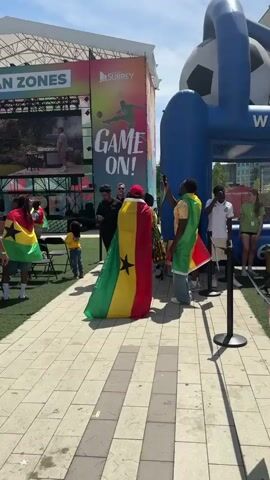 This media captures a lively outdoor event, likely a soccer fan zone, with a crowd gathered under a bright, sunny sky, suggesting it's daytime. The scene is set in Surrey, Canada, indicated by visible signage.

The primary subjects are people, many of whom are enthusiastically displaying their support for Ghana. Several individuals are draped in the Ghanaian flag, which is red, yellow, and green with a black star in the center. Others wear yellow t-shirts, some with names like "NANCY" and "ENOCH" on the back, further emphasizing a connection to the Ghanaian team or community. The attendees appear to be a mix of ages, with a strong sense of collective excitement and pride.

Dominating the background is a large, colorful stage setup with a prominent screen displaying graphics related to soccer, including a silhouette of a player in action and the words "GAME ON!". The word "SURREY" is also visible on the branding of the event. Adjacent to the stage is a large, inflatable soccer goal structure, adding to the sporting atmosphere. In the distance, modern buildings and trees are visible, providing a backdrop of an urban environment.

The activity appears to be a pre-game gathering or a viewing party for a soccer match. The energy is palpable, with people interacting, taking photos, and seemingly anticipating the event. The overall mood is festive and celebratory. The tiled walkway and artificial turf ground indicate a well-organized public space designed for events.